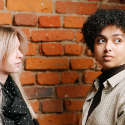 Two people stand in front of a brick wall, one with blonde hair and a black polka dot scarf, the other with curly dark hair and a light-colored shirt.