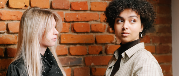 Two people stand in front of a brick wall, one with blonde hair and a black polka dot scarf, the other with curly dark hair and a light-colored shirt.
