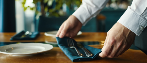 Caucasian young adult man arranging cutlery and napkin on restaurant table, hands visible placing utensils beside empty plates, preparing dining setup for guests